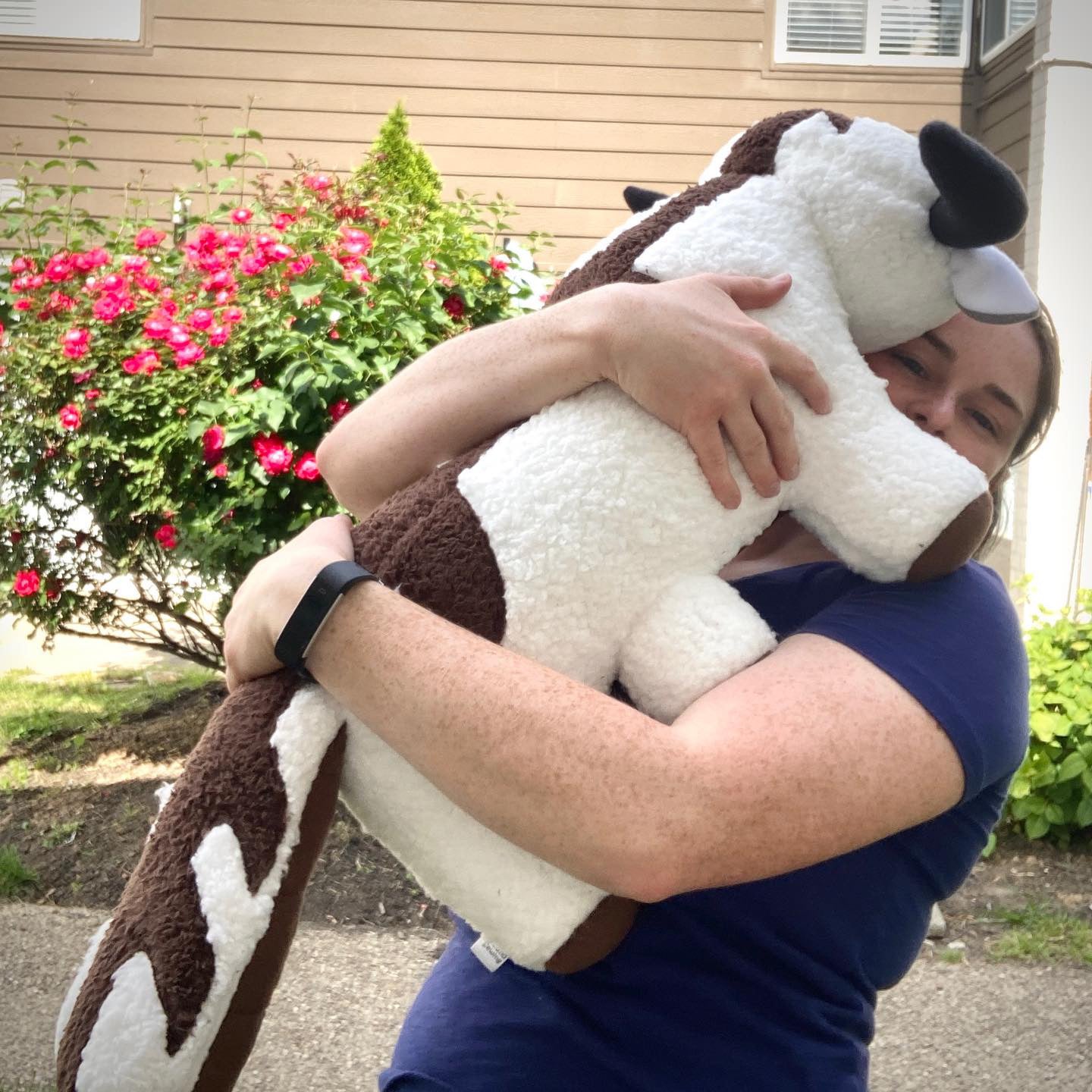 Person holding a large stuffed bison cow toy outdoors with flowers and a house in the background